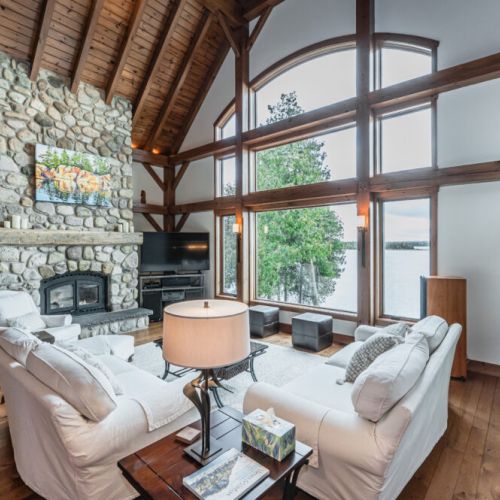 Elegant tranquility meets modern comfort in this spacious post and beam living room, where a majestic stone fireplace and panoramic lake views create an inviting retreat.