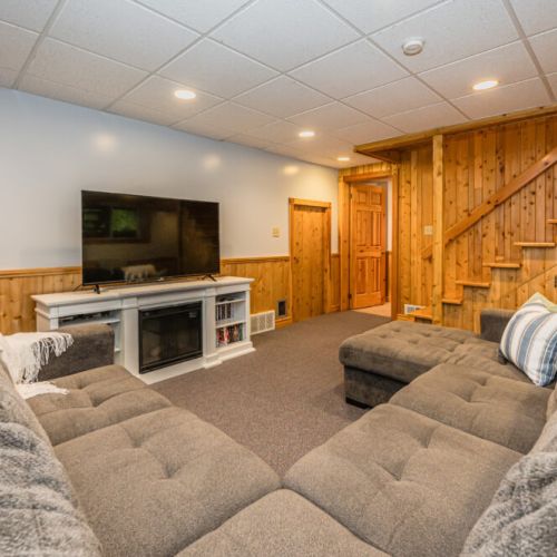 The basement family room features a spacious sectional couch and large TV with electric fireplace  Perfect for movie night!