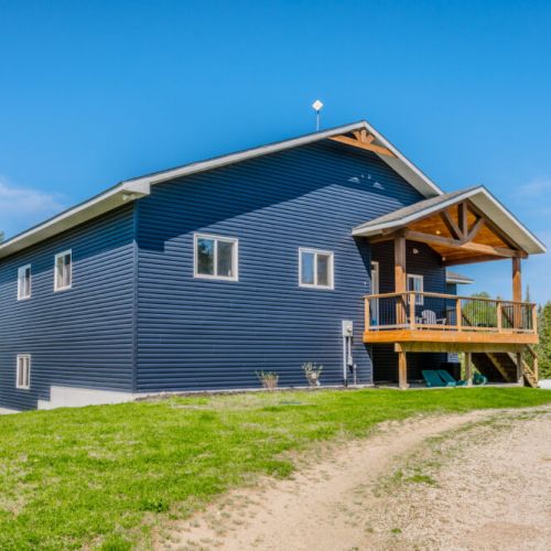 The stunning Charmgri-la cottage, featuring modern blue siding and a rustic wooden porch, stands proudly under a clear blue sky. Surrounded by lush greenery, the cottage offers a perfect blend of contemporary design and natural beauty.