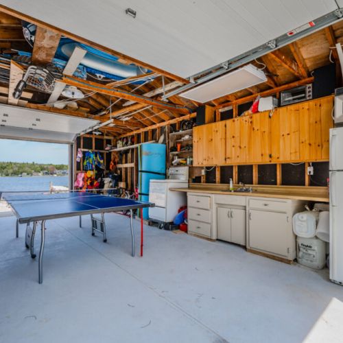 The 'Boathouse' features well-organized storage solutions, including cabinets, shelves, potable water, and a refrigerator. The large garage doors open up to the outdoors, making it easy to transition from indoor activities to lakeside adventures.