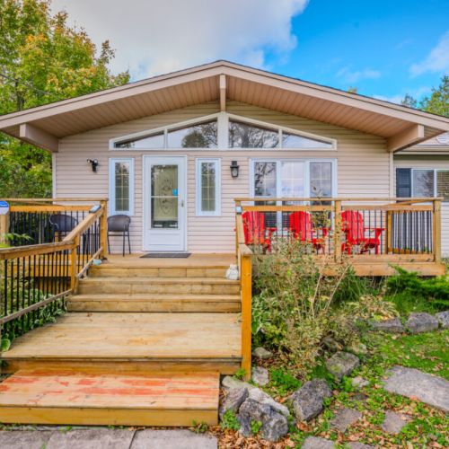 This charming cottage features a welcoming wooden deck with seating, nestled amidst lush greenery and natural stone accents. The modern exterior, highlighted by large windows, offers an inviting space for relaxation and enjoying the outdoors. Perfect for a peaceful retreat in a serene, natural setting.