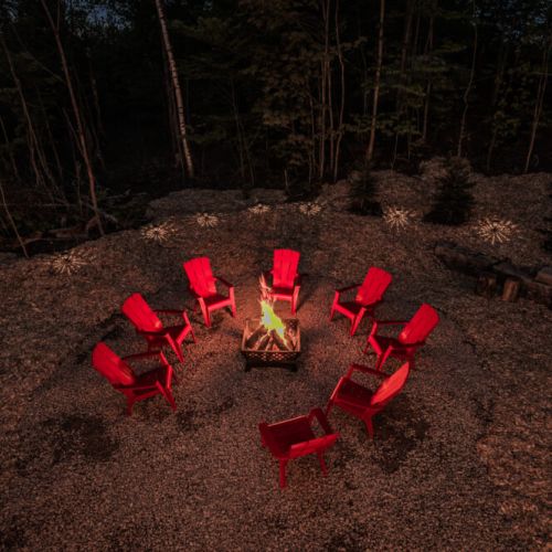The fire pit area is surrounded by red Adirondack chairs, perfect for gathering around the fire in the evening. The setting is enhanced by the soft glow of surrounding lights.