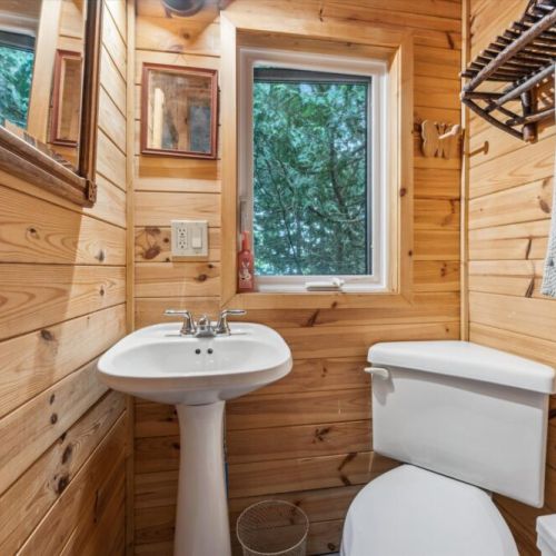 A second bathroom offers cottage comfort with hand-built shelving, a pedestal sink, and calming forest views through the window — all framed in knotty pine.
