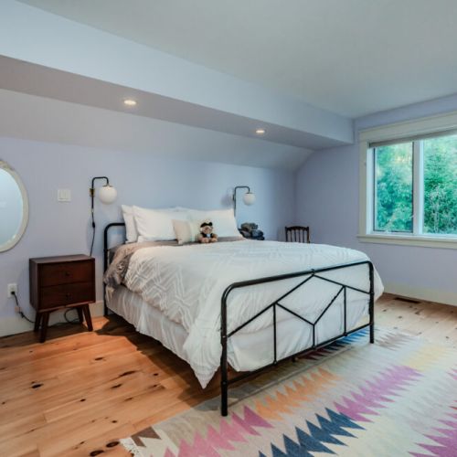 Charming bedroom with soft pastel walls, modern lighting, and a cozy bed, complemented by a vibrant rug—creating a warm and inviting space. (Queen Bed)