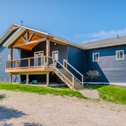 A front view of the spacious Charmgri-la cottage showcases the inviting wooden deck and grand entrance. The expansive gravel driveway provides ample parking space, leading up to this picturesque retreat nestled in the Bruce Peninsula.
