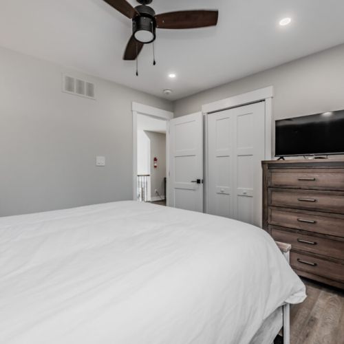 A ceiling fan, recessed lighting, a wooden dresser, and a flat-screen TV provide a comfortable environment for rest. The neutral decor and clean lines create a modern, serene atmosphere, with a double-door closet for extra storage.