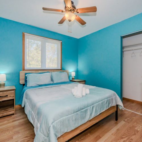 This serene bedroom offers a peaceful retreat, highlighted by a large window that fills the space with natural light. The room is furnished with a comfortable bed, matching nightstands, and a ceiling fan to keep you cool during warm nights. The built-in closet provides plenty of storage space for longer stays.
