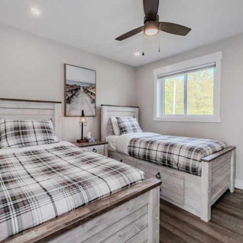 This bedroom features two twin beds with plaid bedding and wooden headboards. The room is bright and airy, with a large window and minimalist decor.