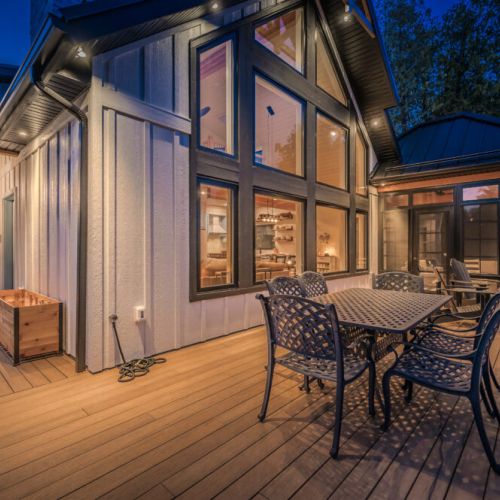 Enjoy serene evenings on the deck of this luxury vacation rental, complete with outdoor dining, ambient lighting, and large picture windows showcasing a cozy, modern interior—perfect for a relaxing retreat near nature.