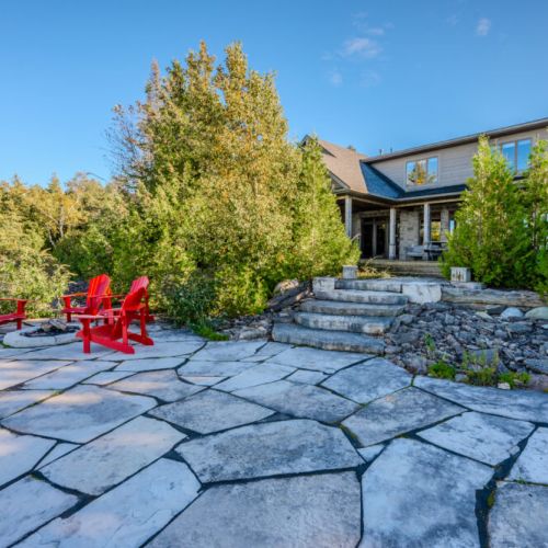 A spacious stone patio with vibrant red  chairs surrounding a fire pit, offering a perfect outdoor retreat with a view of the beautiful house and surrounding greenery.