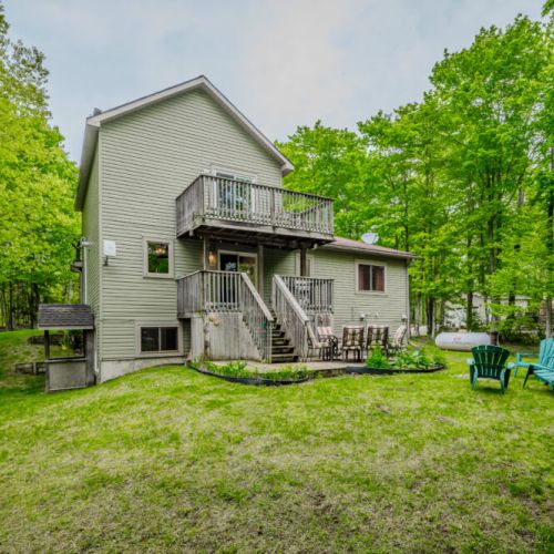 The back view of the cottage showcases a welcoming deck area and ample green space, ideal for outdoor activities and relaxation amidst the lush greenery.