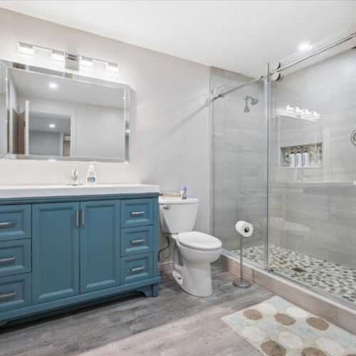 Stylish space featuring a bold blue vanity, large mirrored light fixture, walk-in glass shower with tiled surround, and sleek finishes throughout.