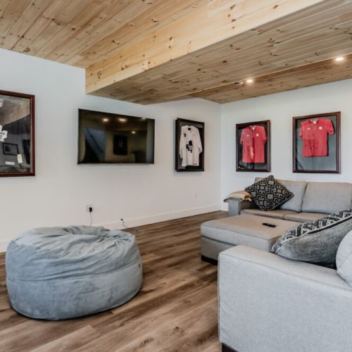 Unwind in our cozy sitting area, featuring a large TV, plush grey sectional sofa, and a bean bag chair. The room is beautifully decorated with framed sports jerseys and a charming wooden ceiling, perfect for relaxing after a day of outdoor adventures.