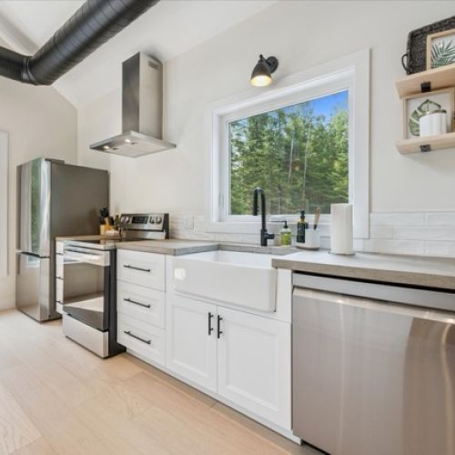 A modern farmhouse kitchen with forest views — featuring stainless steel appliances, open shelving, and a deep apron-front sink for both beauty and function.