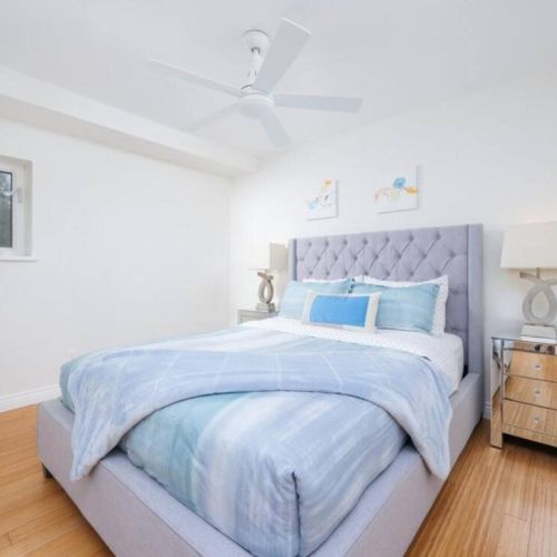 A bright and modern bedroom featuring a tufted upholstered bed with blue bedding, mirrored nightstands, sleek table lamps, and a ceiling fan, all complemented by clean white walls and hardwood flooring.