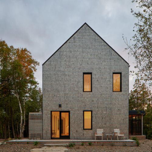 The cottage facade in warm lighting, highlighting the minimalist, wood-shingle architecture against a backdrop of evening skies.