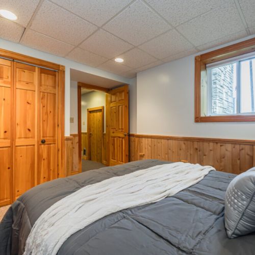 This inviting bedroom features natural wood accents, including paneled closet doors and trim, paired with soft lighting and a cozy window view, creating a warm and serene atmosphere.