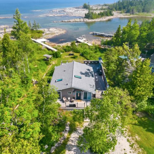 An aerial view reveals a private waterfront cottage surrounded by lush greenery and crystal-clear water. A prime summer travel destination offering privacy, nature, and direct lake access.