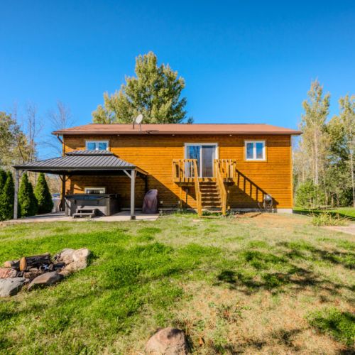 A spacious backyard with a covered patio, a modern grill, and a fire pit. The green lawn is bordered by trees, providing a peaceful, natural setting, perfect for outdoor relaxation and entertaining.