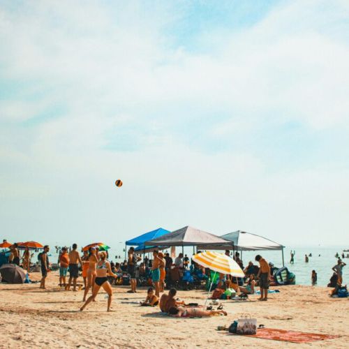 This vibrant beach scene is teeming with the joyful energy of summer. At Sauble Beach, the golden sands serve as a stage for a variety of beachgoers engaged in an array of activities. Families and friends have set up colorful umbrellas and canopies, providing a mosaic of shade from the bright sun. People are lounging on beach towels and chairs, while others engage in playful pursuits like volleyball, with a ball captured mid-air, hinting at the action.

There’s a sense of casual relaxation as some beachgoers wade into the calm, azure waters, while others are content to bask in the sun. A bird is soaring in the clear sky, perhaps on the lookout for a snack or simply enjoying the day. The beach is alive with conversation, laughter, and the carefree ambiance that only a day on the sand by the water can provide. This is a snapshot of summer bliss, a day well-spent where the worries of the world are as distant as the horizon.
