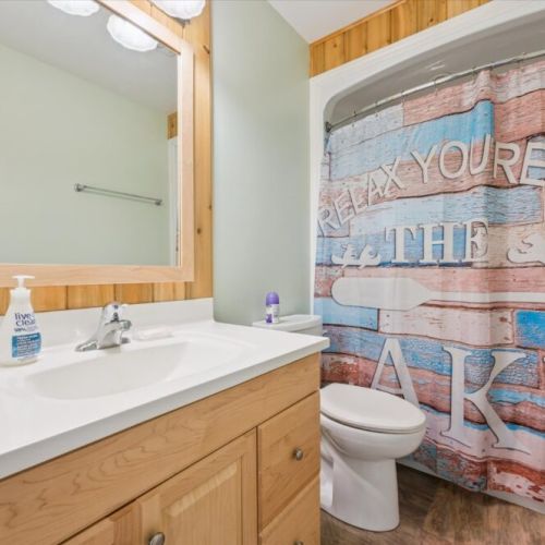 Full bathroom with rustic charm, featuring a light wood vanity, wood-accented mirror frame, and a playful lake-themed shower curtain that reads “Relax, you’re at the lake.”