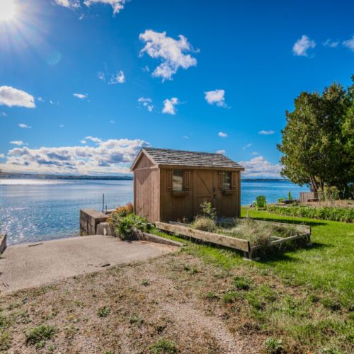 A charming shed sits near the water’s edge, surrounded by lush greenery and a garden space.