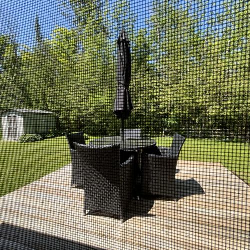 An outdoor patio with wicker furniture and an umbrella. The patio overlooks a lush green yard with two small sheds, surrounded by tall trees, creating a serene and private outdoor space.
