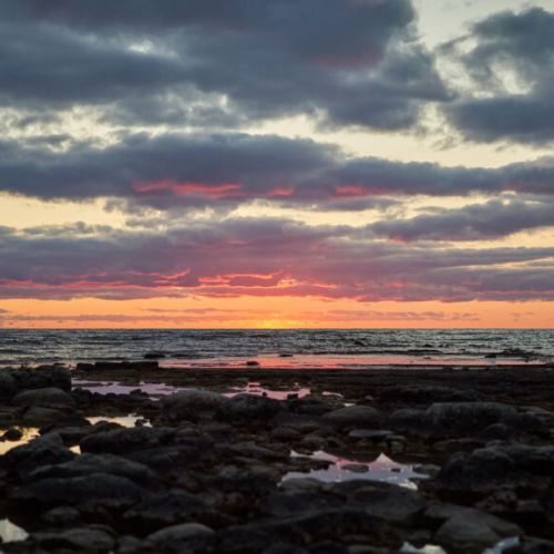 A breathtaking sunset over the water, with reflections on the rocky shoreline creating a dramatic and peaceful scene.