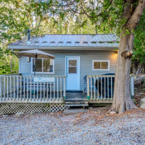 The Oahu: A cozy cabin nestled among tall trees, featuring a welcoming deck with a large umbrella and seating area for outdoor relaxation.