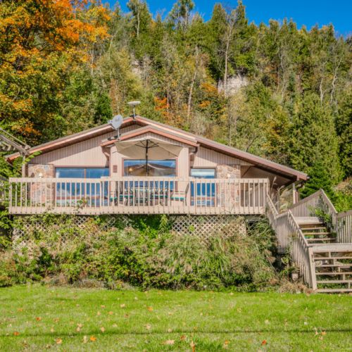 The cottage’s exterior is surrounded by lush greenery, offering a true escape into nature. The spacious deck is perfect for dining al fresco or enjoying morning coffee with a view of the lake.