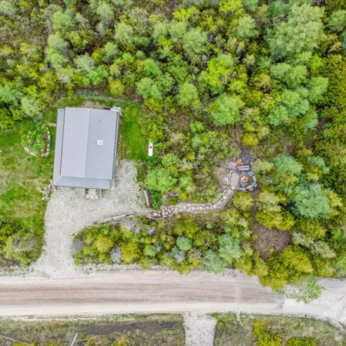 From above, see the scenic layout: gravel drive, wraparound porch, green lawn, and a winding stone path leading to the forest fire pit—every detail designed for both relaxation and exploration.
