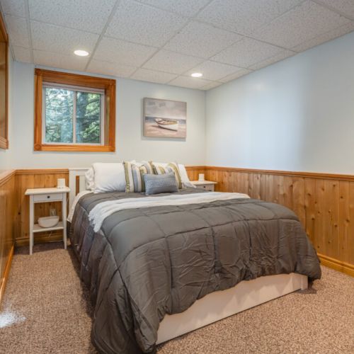 This cozy bedroom features warm wood paneling, a neutral color palette, and a comfortable queen-sized bed adorned with decorative pillows. A large window allows natural light to brighten the space, complemented by recessed lighting.