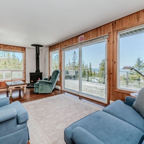 Bright living room with panoramic forest and lake views, cozy seating, and wood stove — the perfect spot to relax after outdoor adventures.