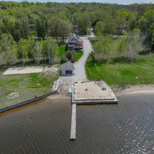 The aerial shot focuses on the beach, dock area and neighbouring lots. The serene lake and surrounding landscape create a perfect backdrop for a relaxing getaway.