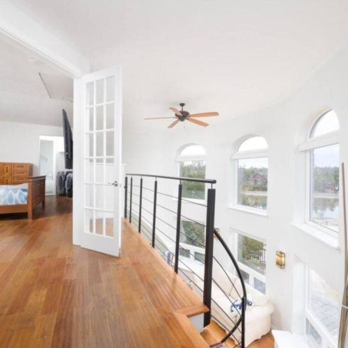 A bright and airy loft space featuring hardwood flooring, a modern railing, and arched windows offering scenic views, leading seamlessly into the master bedroom through elegant French doors.