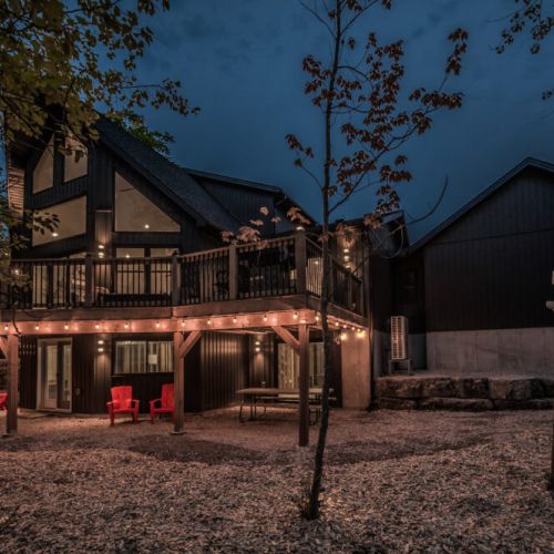 The exterior of the house is beautifully illuminated at night, showcasing the wraparound deck and the surrounding forest. String lights add a cozy ambiance, perfect for evening gatherings.