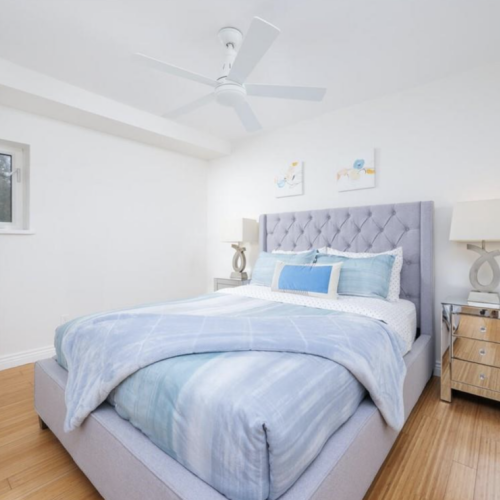 A bright and modern bedroom featuring a tufted upholstered bed with blue bedding, mirrored nightstands, sleek table lamps, and a ceiling fan, all complemented by clean white walls and hardwood flooring.