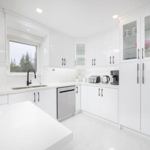 A sleek and contemporary kitchen featuring glossy white cabinetry, quartz countertops, stainless steel appliances, and a black faucet, enhanced by under-cabinet lighting and large windows for natural light.