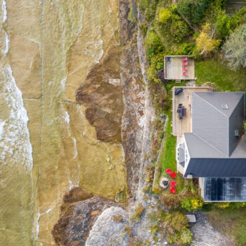 Sure! Here's a more concise version:

---

Aerial view of a modern waterfront home with a spacious deck, fire pit area, and lush greenery, set along a rocky shoreline with clear, shallow waters.