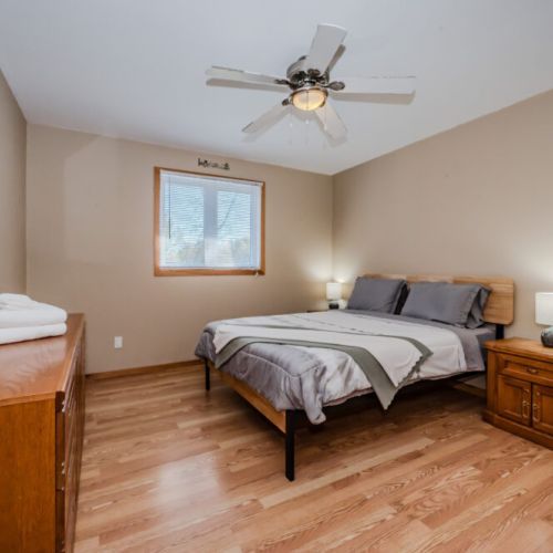 A larger bedroom with simple yet elegant décor, designed for a restful night’s sleep. The room includes a comfortable bed, nightstands with lamps, and a ceiling fan to provide extra comfort. The neutral tones and minimalist furnishings create a calm and peaceful atmosphere for unwinding after a long day.