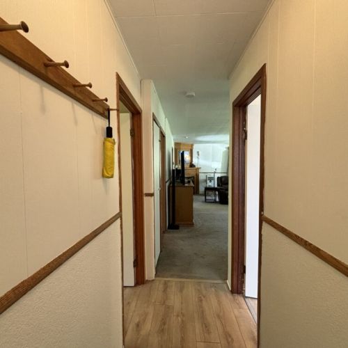 A hallway with neutral walls and wood trim, leading to a living area. A wooden coat rack. The hallway provides access to multiple rooms and opens up into a spacious living area creating a welcoming pathway through the home.