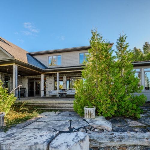 A view of the back of the house showcasing its stone and wood exterior, spacious porch, and surrounded by lush greenery, offering a peaceful and private outdoor space.