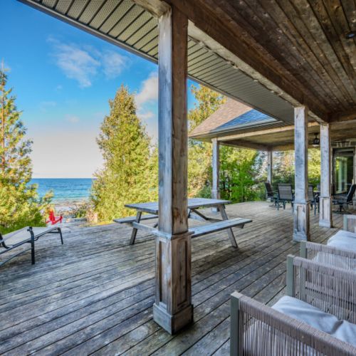 A cozy outdoor area featuring a large covered patio with a picnic table, lounge chairs, and lush greenery surrounding the shoreline.
