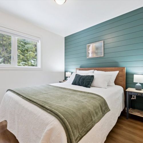 Bright primary bedroom featuring a calming accent wall, warm wood details, and peaceful views of the surrounding woods.