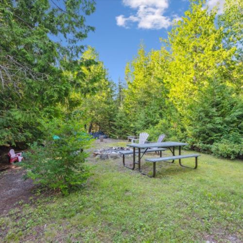 Open grassy area with picnic table and firepit seating, framed by trees — designed for family fun and late-night campfires.