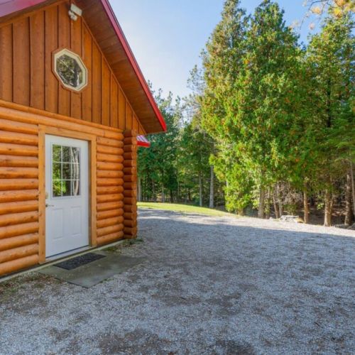 The quaint and discreet main entrance with EV charger to the North side, showcasing the solid craftsmanship of log construction against a backdrop of lush Tobermory forest.