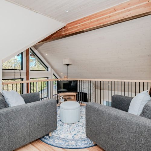 Cozy loft lounge with modern seating and scenic views through large A-frame windows—an ideal spot for relaxation, reading, or streaming during your peaceful cabin getaway.