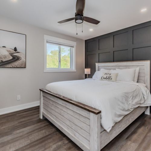This bedroom features a queen-sized bed with white bedding, a dark accent wall, and modern furnishings. The room is brightened by a large window and contemporary decor.