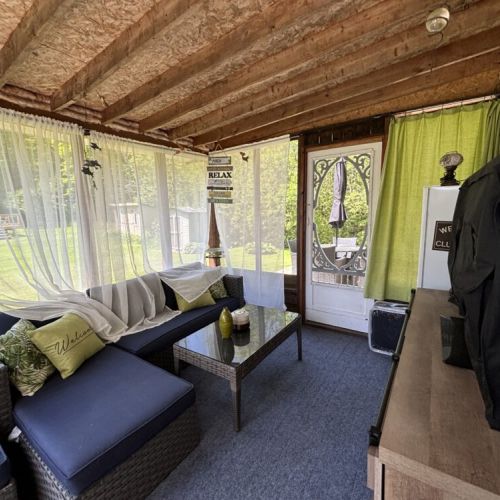 A cozy screened-in porch with sheer white curtains. It features a comfortable wicker sectional sofa with blue cushions and decorative pillows. A glass-top coffee table, adding a functional touch. The porch offers a view of a lush green yard and provides a relaxing space to enjoy the outdoors while staying protected from the elements.