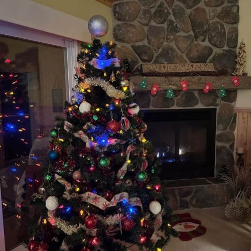 A festive Christmas tree adorned with colourful lights, ornaments, and ribbon, positioned by a cozy stone fireplace. Creating a warm and inviting atmosphere.